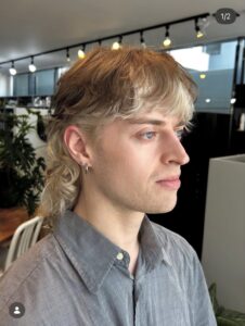 Side profile of a modern shaggy mullet on black hair with a vivid blonde inner color peek-a-boo design, highlighting the layered texture and movement for 2026 hair trends.